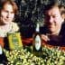 Two individuals posing with bottles of olive oil and a crate of olives in a field. - Olive Oil Times