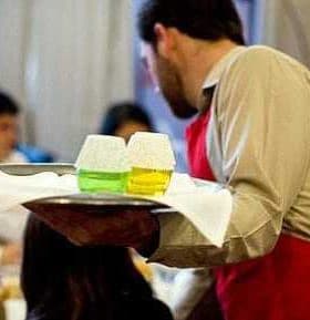 Waiter in a red apron holding a tray with two glasses of colored drinks at a dining event. - Olive Oil Times