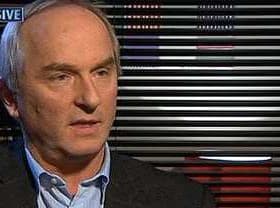 Man with gray hair wearing a dark suit and light shirt during an interview against a striped backdrop. - Olive Oil Times