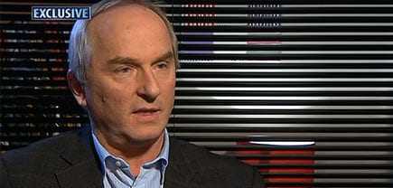 Man with gray hair wearing a dark suit and light shirt during an interview against a striped backdrop. - Olive Oil Times