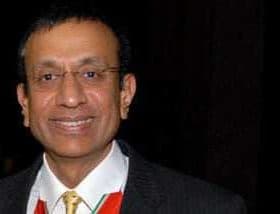 A man wearing a black suit and a tie, smiling at the camera in a formal setting. - Olive Oil Times