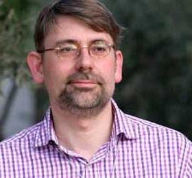 Man wearing a checked shirt with glasses, standing outdoors with a neutral expression. - Olive Oil Times