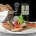 A plate of bruschetta topped with tomatoes and herbs, accompanied by a bottle of olive oil and a basket of bread. - Olive Oil Times