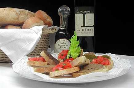 A plate of bruschetta topped with tomatoes and herbs, accompanied by a bottle of olive oil and a basket of bread. - Olive Oil Times