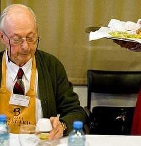 Elderly man wearing glasses and an apron, sitting at a table with a notepad and a bottle of water. - Olive Oil Times