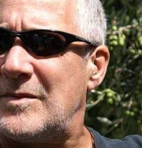 Man with short gray hair wearing sunglasses, with olive trees in the background. - Olive Oil Times