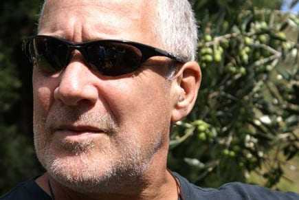 Man with short gray hair wearing sunglasses, with olive trees in the background. - Olive Oil Times