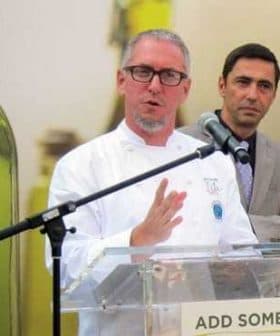 A chef in a white coat speaking at a podium with a microphone during an event. - Olive Oil Times