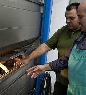 Two men inspecting a baking oven with bread inside, one wearing an apron and the other in a green shirt. - Olive Oil Times