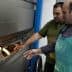 Two men inspecting a baking oven with bread inside, one wearing an apron and the other in a green shirt. - Olive Oil Times