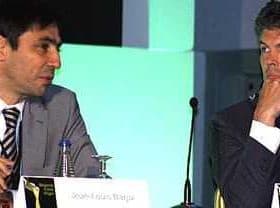 Two men seated at a conference table during a discussion, one wearing a suit and the other in a blazer. - Olive Oil Times