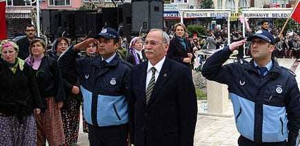 Two police officers saluting alongside a man in a suit at a public event with a crowd in the background. - Olive Oil Times