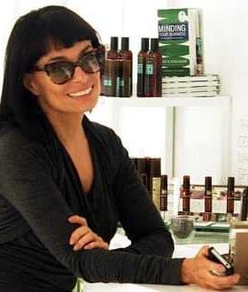 Woman with dark hair and sunglasses smiling while leaning against a counter with products in the background. - Olive Oil Times
