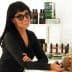 Woman with dark hair and sunglasses smiling while leaning against a counter with products in the background. - Olive Oil Times