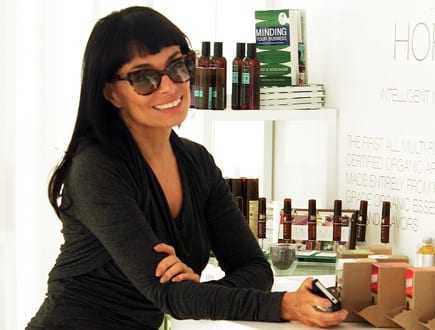 Woman with dark hair and sunglasses smiling while leaning against a counter with products in the background. - Olive Oil Times