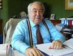 An older man with gray hair wearing a blue shirt and suspenders sitting at a desk covered with papers and files. - Olive Oil Times