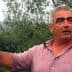 Man with gray hair gesturing while standing in an olive grove with trees in the background. - Olive Oil Times