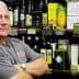 Man with gray hair standing with arms crossed in front of olive oil bottles on shelves. - Olive Oil Times