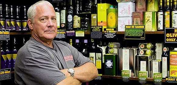Man with gray hair standing with arms crossed in front of olive oil bottles on shelves. - Olive Oil Times