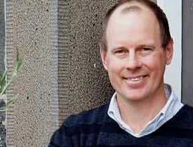 Man with short hair wearing a navy blue sweater, smiling in front of a textured wall. - Olive Oil Times
