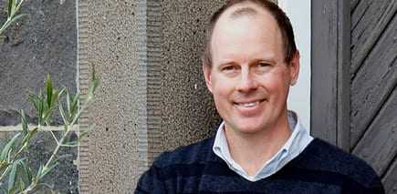 Man with short hair wearing a navy blue sweater, smiling in front of a textured wall. - Olive Oil Times