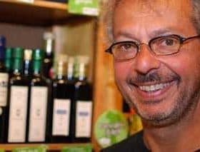 Man with glasses smiling in front of shelves displaying olive oil bottles. - Olive Oil Times