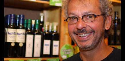 Man with glasses smiling in front of shelves displaying olive oil bottles. - Olive Oil Times