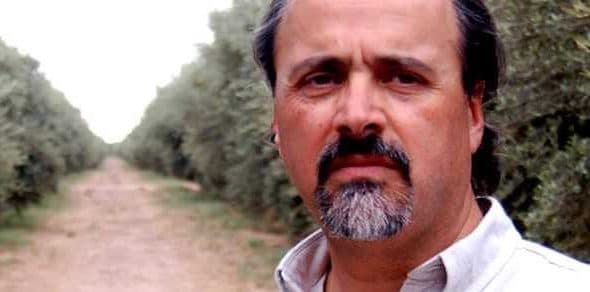 A man standing in an olive grove with trees lining a dirt path in the background. - Olive Oil Times