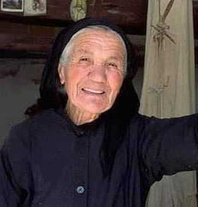 Elderly woman wearing a black dress and headscarf, smiling while leaning against a wall. - Olive Oil Times