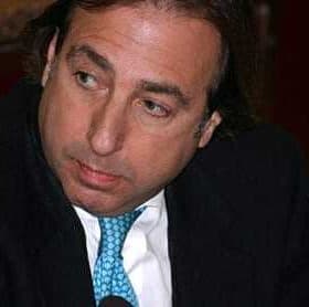 Man wearing a black suit and a blue tie, looking to the side during a discussion. - Olive Oil Times