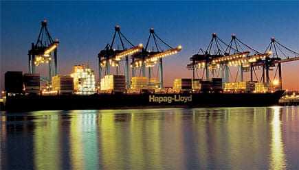 A large container ship named Hapag-Lloyd docked at a port with cranes illuminated at dusk. - Olive Oil Times