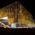 Contemporary building featuring a yellow facade with a textured surface, illuminated at night. - Olive Oil Times
