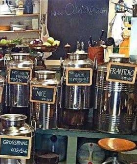 Various stainless steel containers for olive oil displayed on a shelf with labels indicating different types. - Olive Oil Times