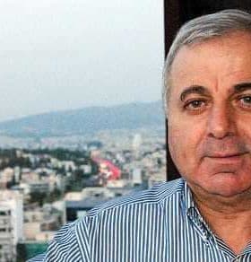 A man with gray hair wearing a striped shirt, posing for a portrait with a cityscape in the background. - Olive Oil Times