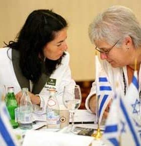 Two individuals seated at a table, engaged in a discussion with various flags and drinks visible. - Olive Oil Times