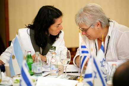 Two individuals seated at a table, engaged in a discussion with various flags and drinks visible. - Olive Oil Times