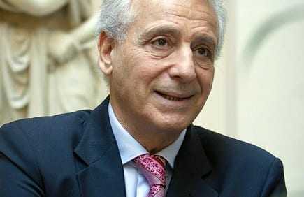 A man wearing a dark suit and a pink tie, smiling while seated indoors. - Olive Oil Times