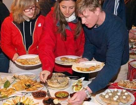 Three individuals selecting food from a buffet table with various dishes and plates. - Olive Oil Times