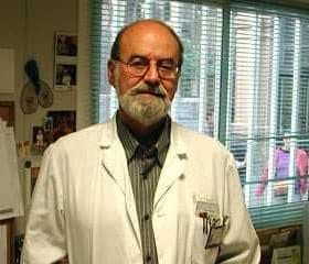 A man wearing a white lab coat stands in an office with a computer and various papers around. - Olive Oil Times