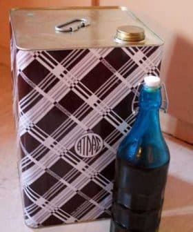 A square metal container with a patterned design next to a blue glass bottle. - Olive Oil Times