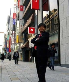 Businessman in a suit standing with arms crossed on a city street with storefronts in the background. - Olive Oil Times