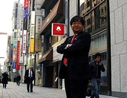 Businessman in a suit standing with arms crossed on a city street with storefronts in the background. - Olive Oil Times