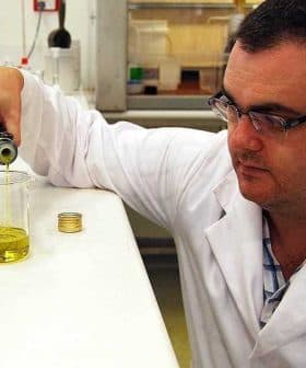 Scientist in a lab coat pouring olive oil from a bottle into a beaker in a laboratory setting. - Olive Oil Times