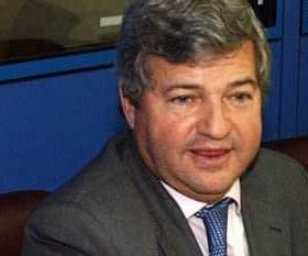 A man wearing a gray suit and blue tie seated at a table during a conference or meeting. - Olive Oil Times