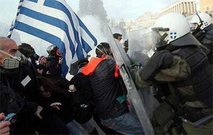 A protest scene in Greece featuring a person holding a Greek flag and police in riot gear. - Olive Oil Times