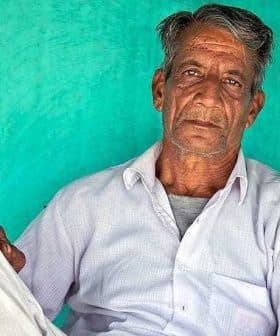An elderly Indian farmer sitting against a turquoise wall, wearing a white shirt and pants. - Olive Oil Times