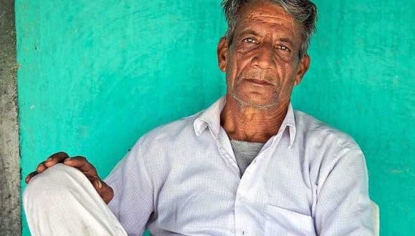 An elderly Indian farmer sitting against a turquoise wall, wearing a white shirt and pants. - Olive Oil Times