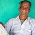 An elderly Indian farmer sitting against a turquoise wall, wearing a white shirt and pants. - Olive Oil Times