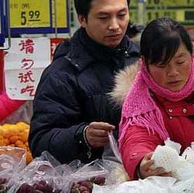 Two individuals selecting fruits in a market with price tags visible in the background. - Olive Oil Times