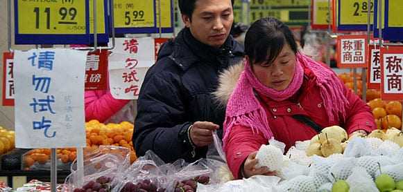 Two individuals selecting fruits in a market with price tags visible in the background. - Olive Oil Times
